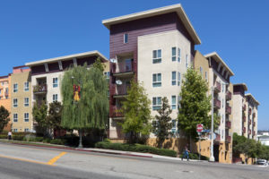 Exterior shot from street corner with balconies, trees, and people walking on sidewalk