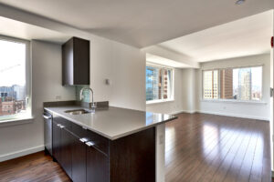 Unit living room/Kitchen, Caesarstone countertop, breakfast nook parallel to sink, dark brown cabinetry, stainless steel appliances, hardwood floors, white walls, windows on each wall from left to right in photo. Photo taken in kitchen lookout out towards living room.