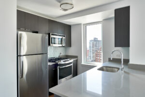 Unit Kitchen, stainless steel appliances, dark wood cabinetry, white walls, green subway tile from cabinets to countertops, sink attached to kitchen island, breakfast nook parallel to sink.