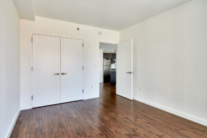 Unit Bedroom, hardwood floors, white walls, closet doors, photo taken inside bedroom looking out to kitchen/living room.