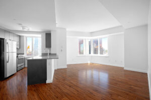Unit kitchen/Living room, breakfast bar kitchen countertop, open floorplan, hardwood floors, stainless steel appliances, windows on both sides of corner in the living room, white walls.
