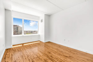 Unit Bedroom, hardwood floors, large wall to wall windows on far side wall, reading nook area in front of window, white walls.