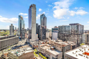 Unit view of city underneath, sprawling skyline, buildings in distance, photo taken on a sunny day with some clouds.