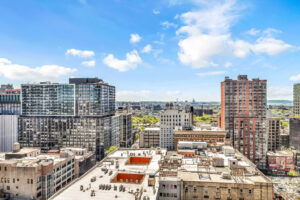 Unit view of city underneath, sprawling skyline, buildings in distance, photo taken on a sunny day with some clouds.