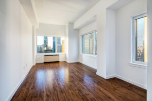 Unit Living room, windows on each far side wall, 3 total in view, furnace/ac along end of living room in front of window.
