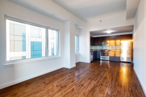 Unit Living room, windows running alongside left side of room, photo taken at the end of the living room towards the kitchen, hardwood floors, Stainless steel appliances, white walls.