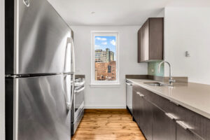 Alternate Unit Kitchen, Window with view of city, stainless steel appliances, dark brown cabinetry, hardwood floors, Breakfast bar, sink, dishwasher, green Subway tile, white walls.