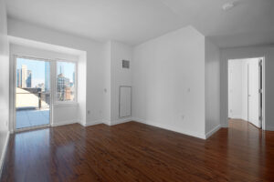 Unit Living room, open floorplan, window and door leading to patio, hardwood floors, white walls, hallway door to bathrooms and bedrooms.