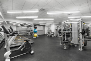 Community Fitness room alternate view, white walls, gray matted carpet, stationary bicycle, treadmills, mirrored walls, weightlifting section, free weight section, yoga balls.