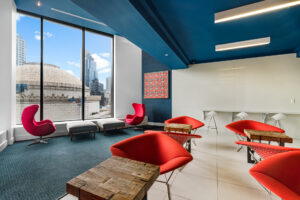 Community Lounge Room alternate view, 4-2 chair tables, bar seating along perimeter, kitchen sink, TV, Tile and Carpeted Floor, Square contemporary art on white & blue walls, floor to ceiling windows with view of city, seating near window.