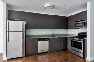 Unit Kitchen/Living room, open floor plan, hardwood floors, dark wooden cabinetry, stainless steel appliances, green subway tile between cabinetry and countertop.