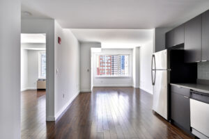 Unit Kitchen/Living room, open floor plan, hardwood floors, stainless steel appliances, dark brown cabinetry, window on far side living room wall, photo taken from kitchen looking into living room and bedroom (on left side of photo).