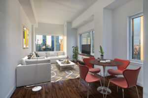 Unit Living room, from right to left, 4 seat contemporary dining table, window on wall parallel to camera and right-side wall, entertainment center with tv on top, sectional couch parallel to far side window and entertainment center, large contemporary area rug, Hardwood floors, potted plants in corners with windows, marble coffee table between couch and tv.