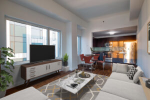 Unit Living room, from left to right, entertainment center with tv atop and window behind tv, large sectional couch pointed towards kitchen area and tv, marble coffee table between couch and tv, large contemporary area rug beneath couch and coffee table, potted plant in left hand corner of room, 4 seat dining table between living area and kitchen, wooden floors, stainless steel appliances, dark brown wooden cabinetry in kitchen, sunlight cast on kitchen area from windows.