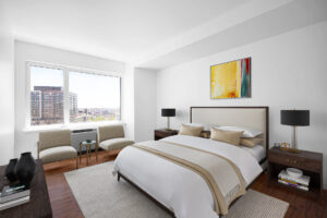 Unit Bedroom, Furnished, king size bed, abstract art hung above bedframe, wooden nightstands on both sides of bed, rattan-like rug beneath bed, 2 reading chairs in front of window on left side of photo, dresser and tv parallel to bed, hardwood floors, white walls.