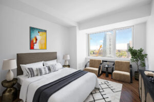 Unit Bedroom, Furnished, King size bed, Abstract art hung above bedframe, contemporary brass nightstands on both sides of bed, dresser parallel to bed. Lounge/reading chairs placed in front of window on far side wall, Potted plant in corner next to window and chairs.