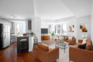 Unit Kitchen/Living room, Furnished, from left to right; Kitchen area, stainless steel appliances, sink on kitchen island, breakfast nook parallel to sink, dark wooden cabinetry, window between oven and sink, hardwood floors, Middle of photo, couch and seating area right of breakfast nook, glass coffee table between chairs facing each other and couch facing breakfast nook, Area rug beneath both lounge chairs, couch and coffee table, behind right side lounge chair is a 4 seat glass dining table, to the left of the dining table is the entertainment center and mounted tv above windows along wall to the right of tv, when glass dining table ends walls begin again and hung there are two abstract paintings.