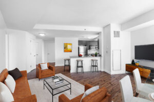 Unit Living/Kitchen room, Furnished, Open floorplan, hardwood floors, white walls, From left to right; Couch and lounge chairs with glass coffee table in-between, large area rug beneath couch, chairs, and coffee table, breakfast nook/ kitchen island right of far side lounge chair, behind is the kitchen area with stainless steel appliances, to the right of the breakfast nook is the entertainment center with a mounted tv hung above, in front of the nearside lounge chair is the 4 seat glass dining table, to the left of the kitchen is a hallway leading to the entranceway/bedrooms/bathrooms.