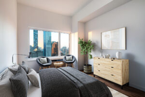 Unit Bedroom, Furnished, left side of room, queen size bed, right side of room, dresser, potted plant left of dresser, two reading chairs with end table in-between left of potted plant, chairs lie in front of window looking out to the city, white walls and contemporary art hung above dresser.