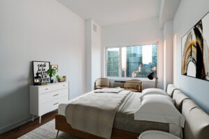 Unit Bedroom, Furnished, queen size bed on right side of room, dresser parallel to bed, nightstands on both side of bed, two chairs in front of window on far side wall of photo, contemporary art hung above bed and place on top of dresser. Neutral colored furniture.