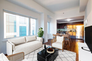 Unit Living room, Left to Right; couch along left side wall, window above couch, lounge chairs parallel to each other on both sides of couch, black marble coffee table in middle of couch and chairs, 4 seat dining table behind right-side lounge chair, Kitchen behind dining table, TV/ entertainment center parallel to couch, white walls, hardwood floors.