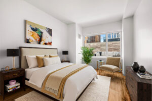 Unit Bedroom, hardwood floors, queen size bed left side of room, dresser parallel to bed, nightstands on both sides of bed, rattan-like rug beneath bed, reading chair in corner by window facing bed, potted plant adjacent to reading chair, abstract art hung above bed.