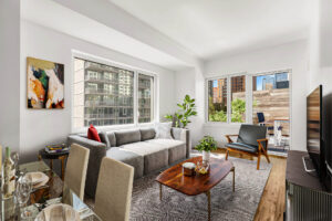 Unit living room, left to right; 4 seat glass dining table, abstract art hung left of large window, end table beneath abstract art, gray couch to right of end table and below window, wooden coffee table in front of couch, black and white geometric rug beneath couch and coffee table, entertainment center with tv atop parallel to couch on other side of the room, reading chair facing towards dining table, window and patio door behind reading chair, outdoor dining area and plants on patio.