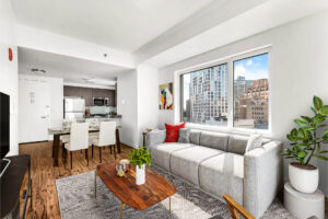 Unit Living room, furnished, From Right to left; potted plant in right hand corner of room, couch left of potted plant, wooden coffee table in front of couch, black and white geometric rug underneath couch and coffee table, entertainment center parallel to couch on left side of room, 4 chair glass dining table to left of couch, end table left of couch, abstract art hung above end table, entryway door behind dining table, kitchen in view right of entryway door.