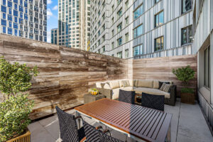 Unit terrace, furnished, 4 seat outdoor dining table, outdoor sectional couch behind dining table, end table left of sectional, potted plant right of sectional, wooden fencing for privacy.