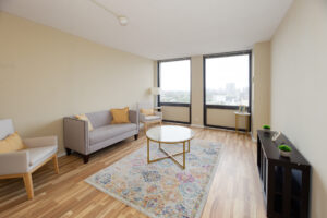 Unit Living room, furnished, 2 large windows, 2 person couch, 2 lounge chairs, large round marble coffee table, multicolored area rug, wood floors.