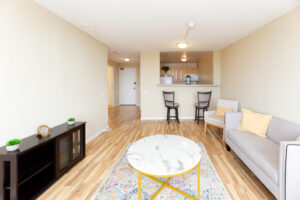 Unit Living room, Breakfast bar joined to kitchen, 2 bar stools, 2 person couch, kitchen attached to living room, multicolored area rug, marble coffee table in center of room.