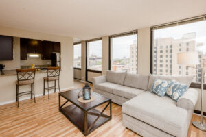 Unit Living Room, Large Sectional Couch, Breakfast bar attached to kitchen, Wood Floors, Brown Coffee table, Windows alongside perimeter.
