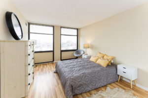 Unit Bedroom, wood floors, queen sized bed, 2 nightstands, chair in corner by windows. 2 large windows with view of downtown.