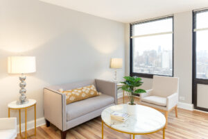 Unit Living room, wood floors, 2 large windows, 2 person couch and chairs placed around marble coffee table.